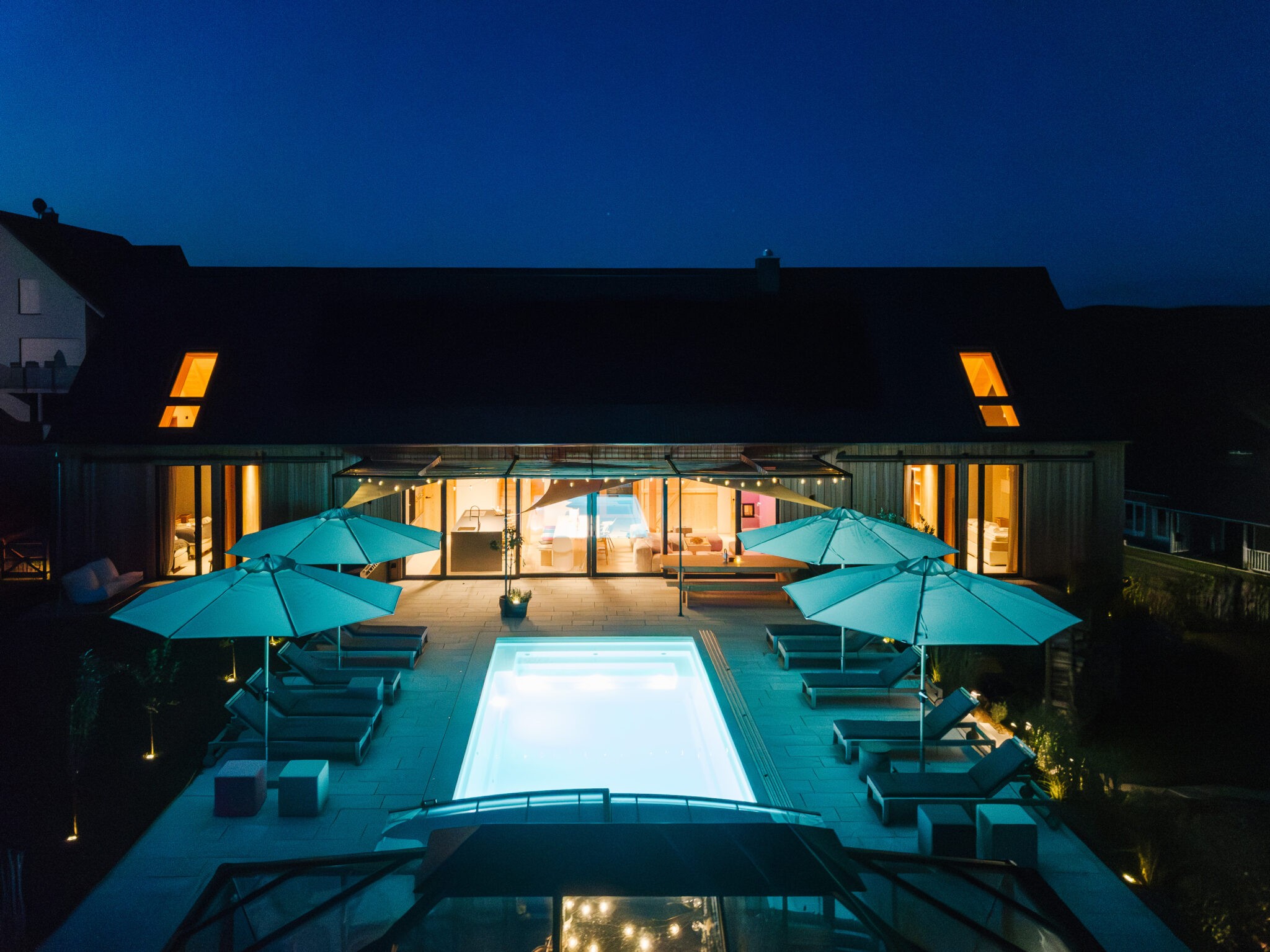 Sichtbar ist ein großes Ferienhaus, verkleidet mit Holz, mit einem beleuchteten Pool bei Abendlicht. 4 Sonnenschirme sind aufgespannt